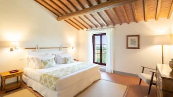 Un dormitorio con techo de madera, una cama con ropa blanca, una mesita de noche, una silla y una ventana con cortinas.