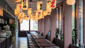 A restaurant with wooden tables and chairs, hanging lanterns with Japanese characters, and potted plants on the windowsill.