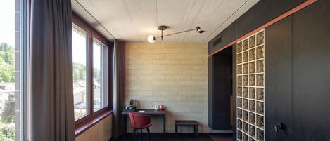 A hotel room with a bed, a desk with a chair, a small table, a window with curtains, and a brick wall with a decorative panel.