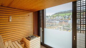 A sauna room with wooden benches, a wooden bench with a bucket, and a window with a city view.