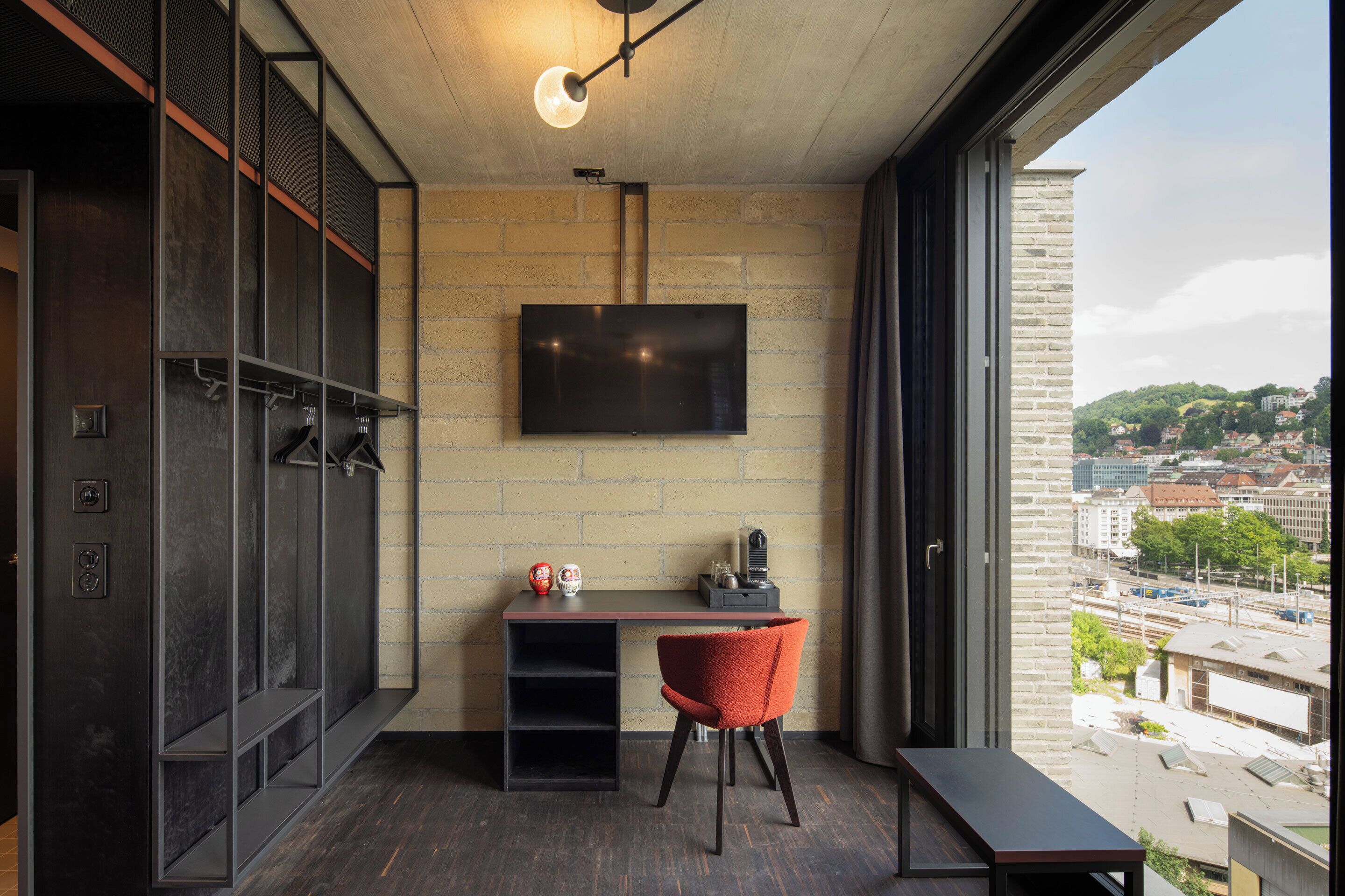 A modern room with a large window, a flat-screen TV mounted on a brick wall, a desk with a chair, and a bench.