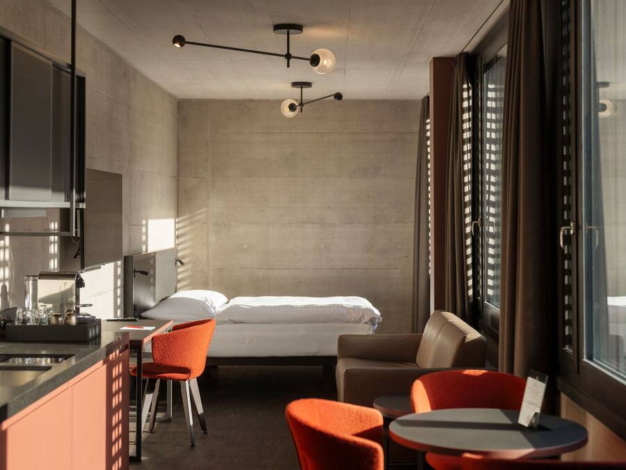 A compact hotel room with a bed, a small kitchenette, a dining area with a table and chairs, and large windows providing natural light.