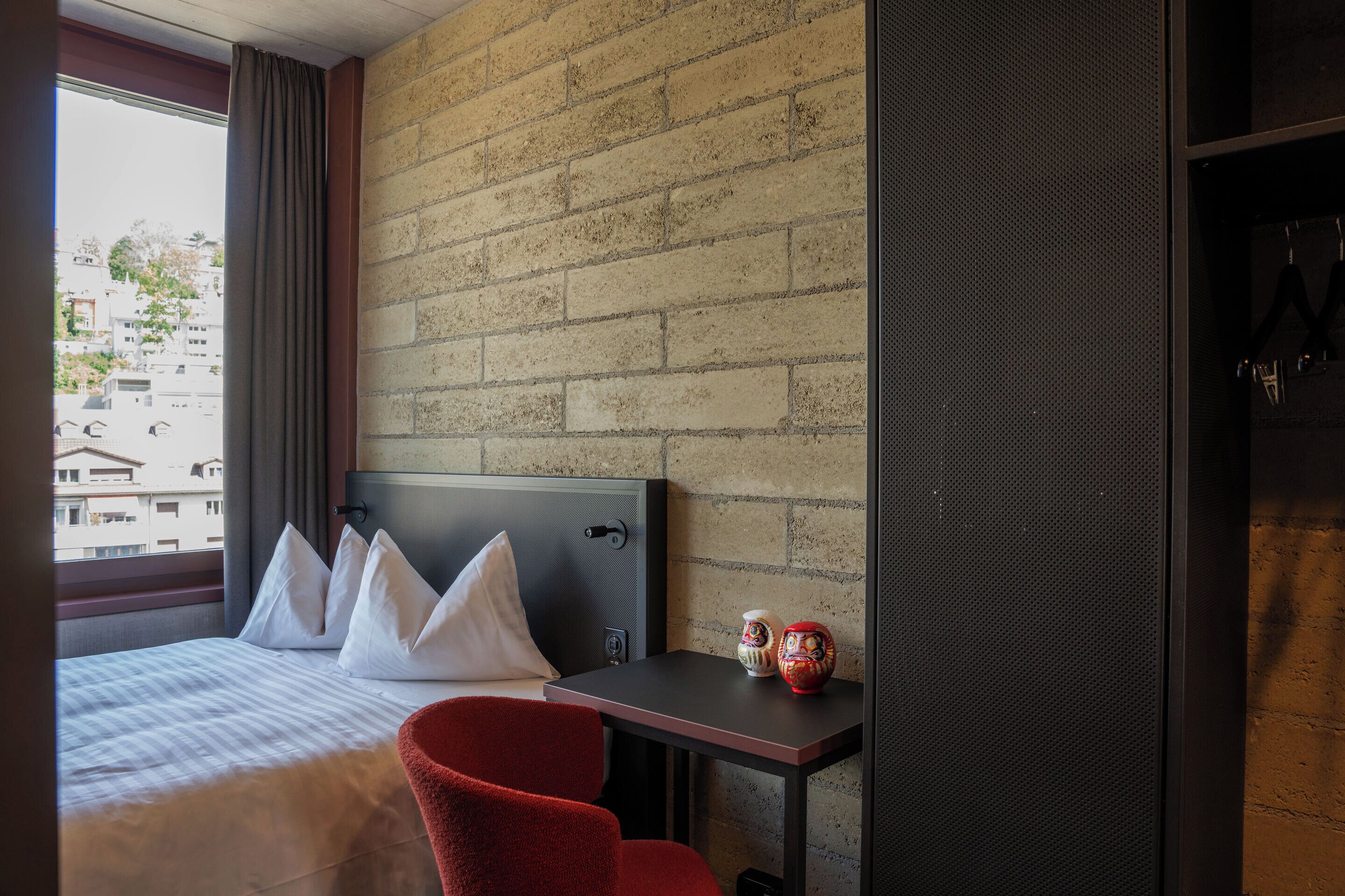 A hotel room with a bed, a red chair, a small table with decorative items, and a brick wall.