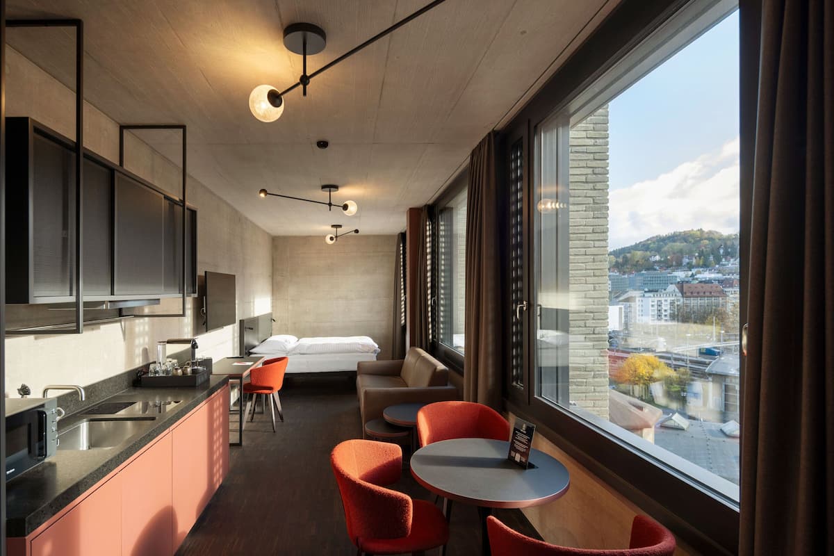 A modern hotel room with a kitchen, a dining area with orange chairs, a bed, and a large window offering a city view.