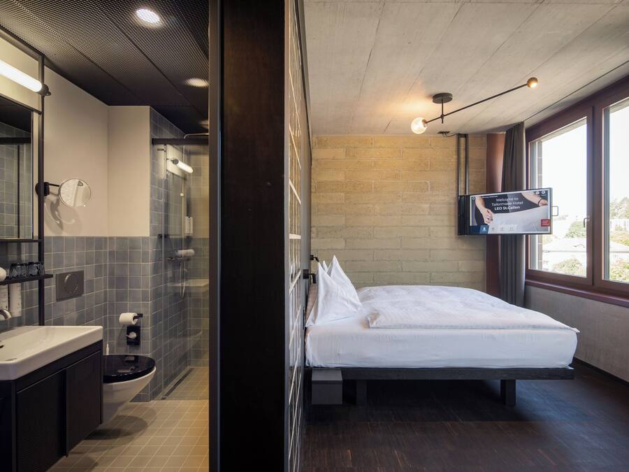 A modern hotel room with a large bed, a bathroom with a shower, and a TV mounted on the wall.
