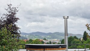 A wooden hot tub on a rooftop with a metal chimney, surrounded by greenery and overlooking a cityscape.