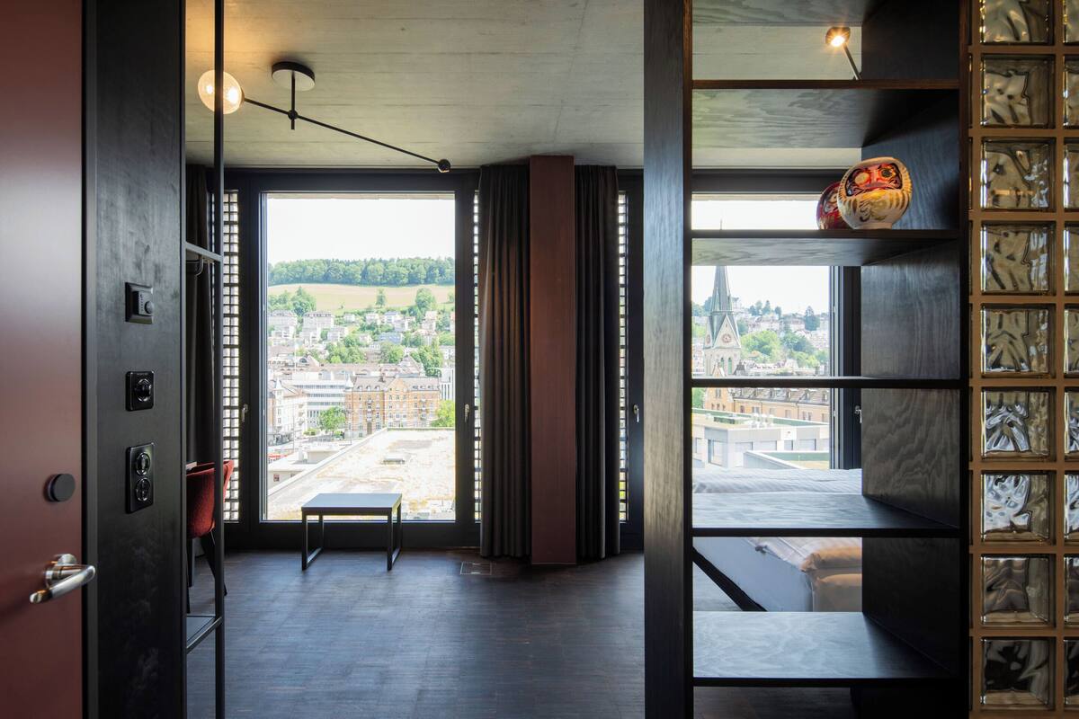 A modern hotel room with a large window, a bench, and a view of the cityscape.