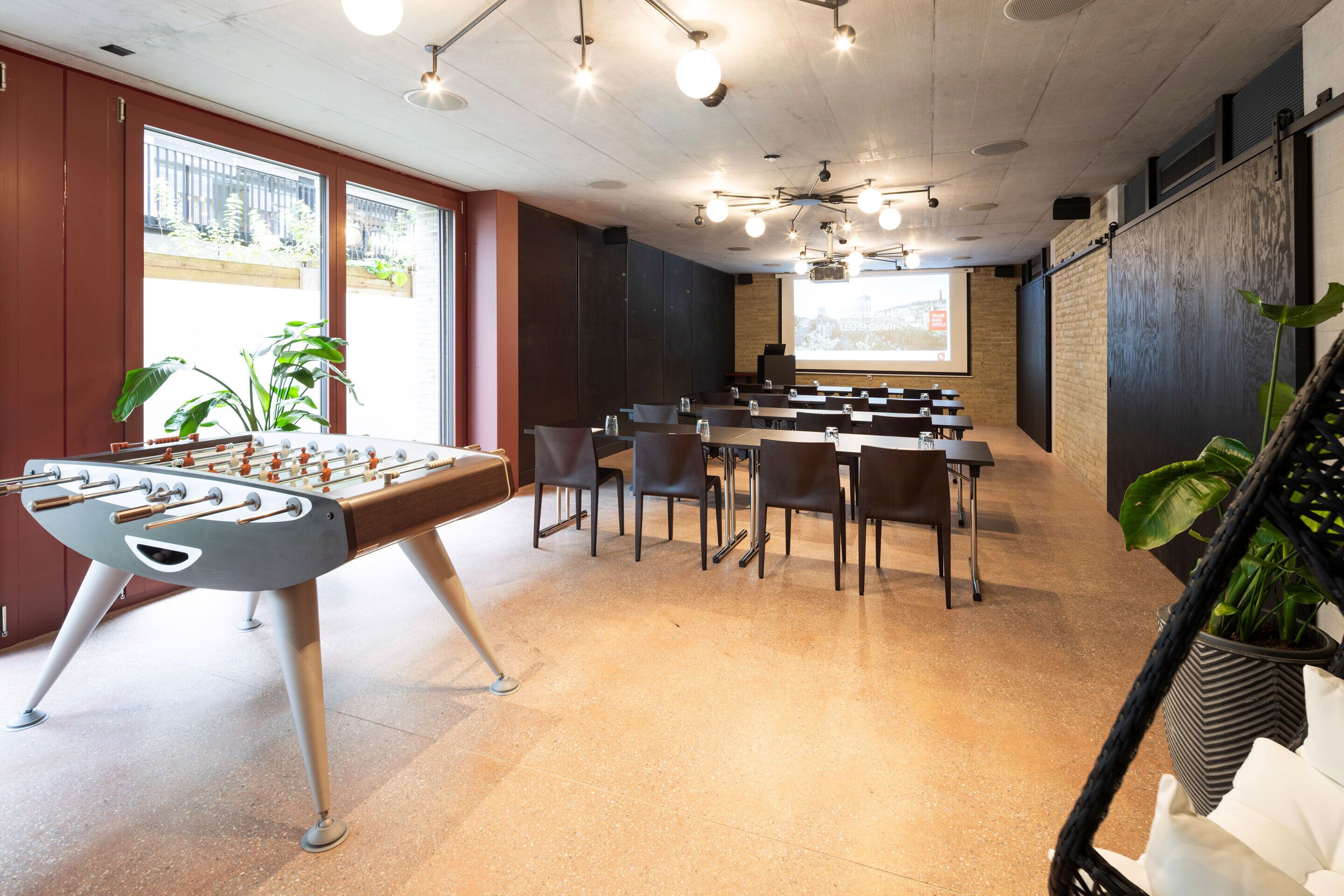 A modern meeting room with a foosball table, several chairs, a projector screen, and large windows with plants.