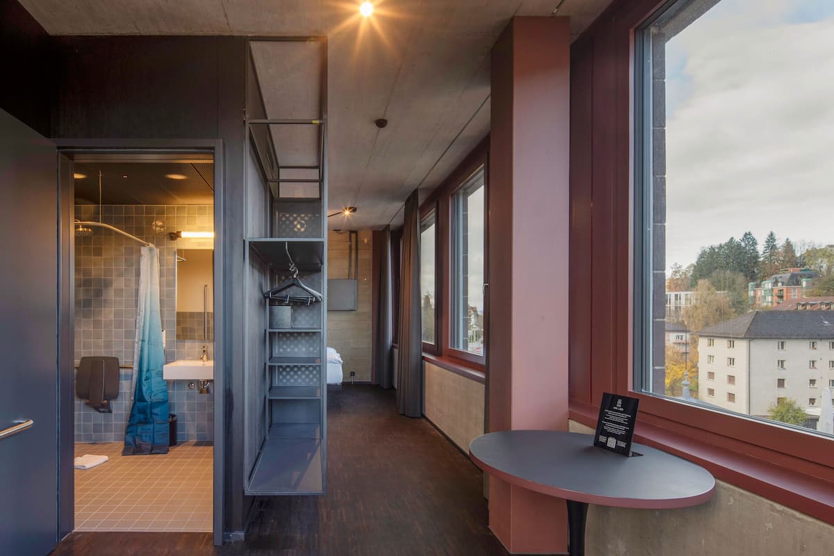 A modern hotel room with a view of the city, a small table with a sign, and a bathroom with a shower and towel rack.