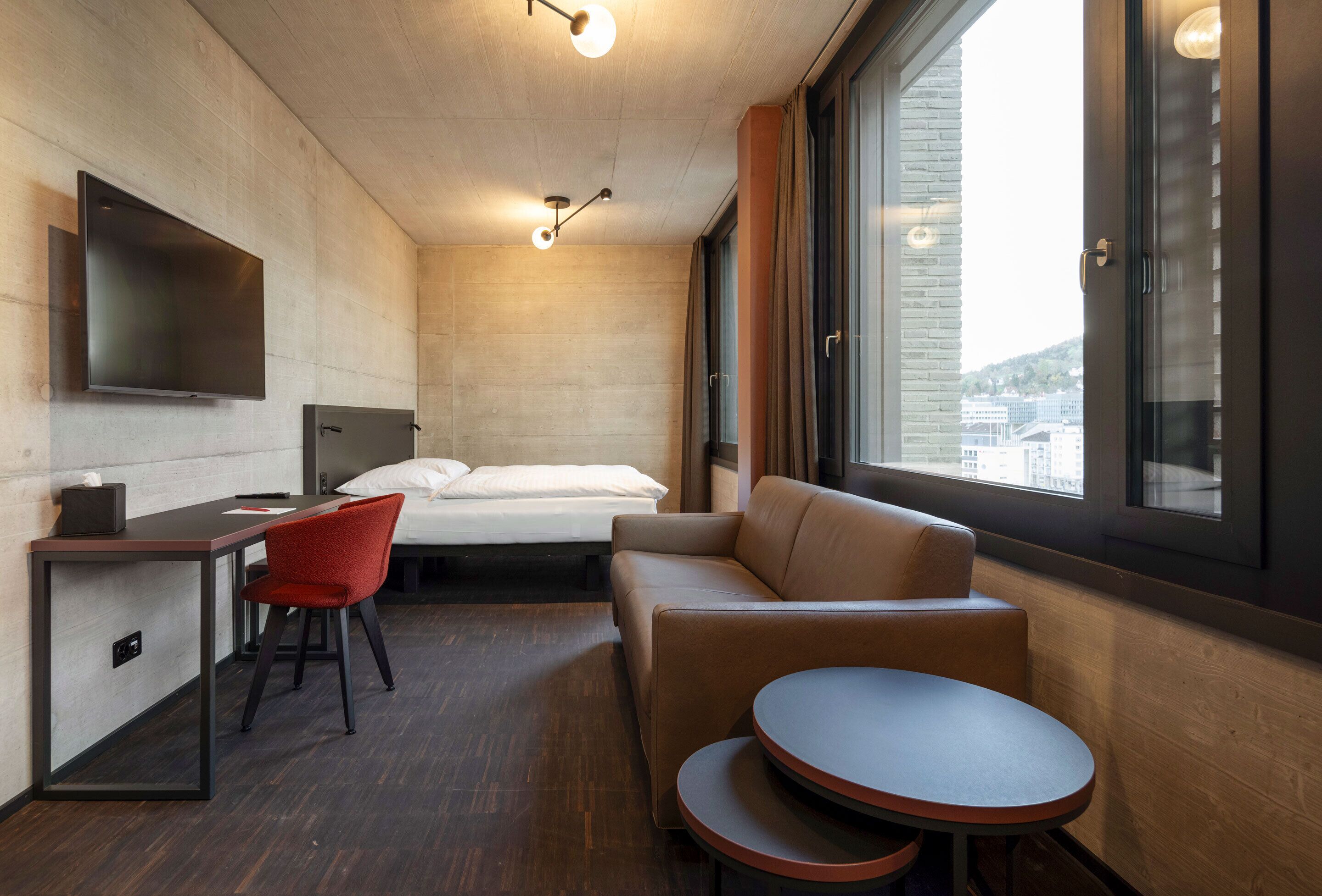 A modern hotel room with a bed, a desk with a chair, a sofa, a small table, and a TV mounted on the wall.
