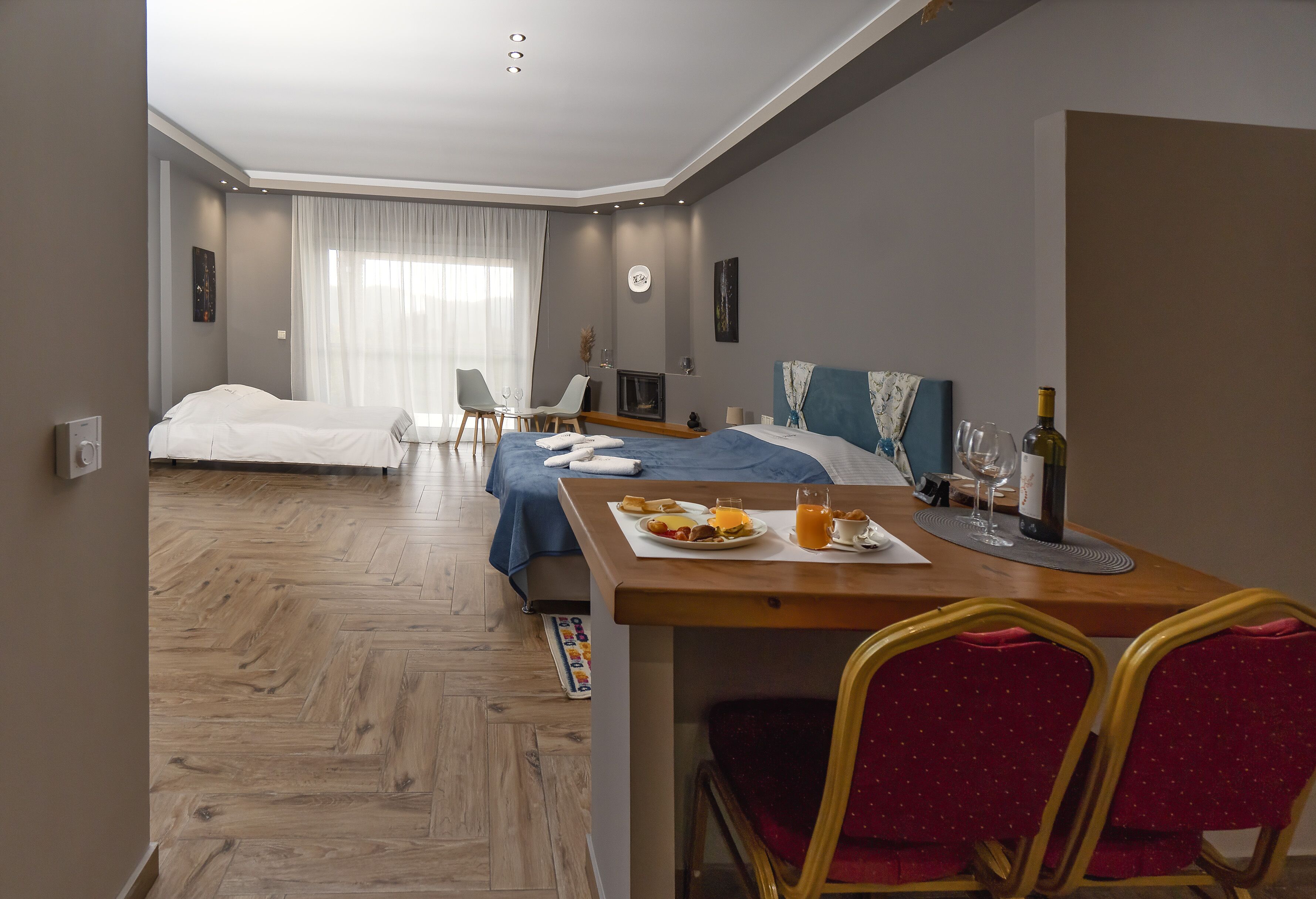 Ein modernes Hotelzimmer mit Holzboden, einem Bett mit blauer Kopfteil, einem Esstisch mit Frühstück und einem Kamin.