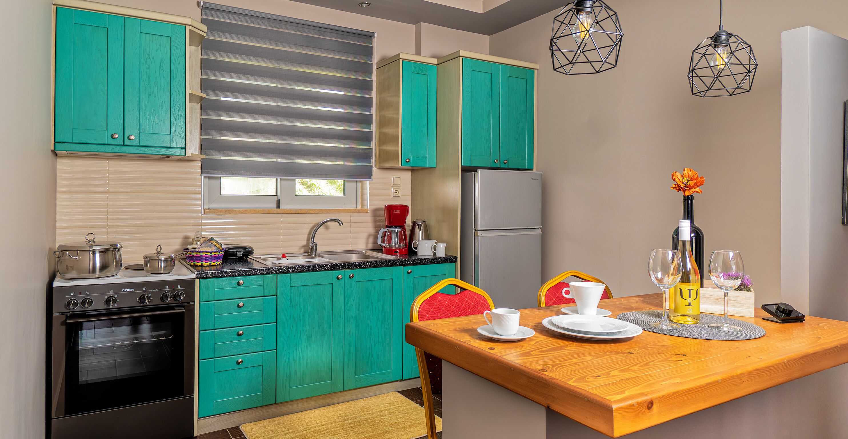 Una cocina con armarios azul verdoso, una estufa y un refrigerador. Hay una mesa de comedor con un mantel individual, una silla roja y un florero con flores.