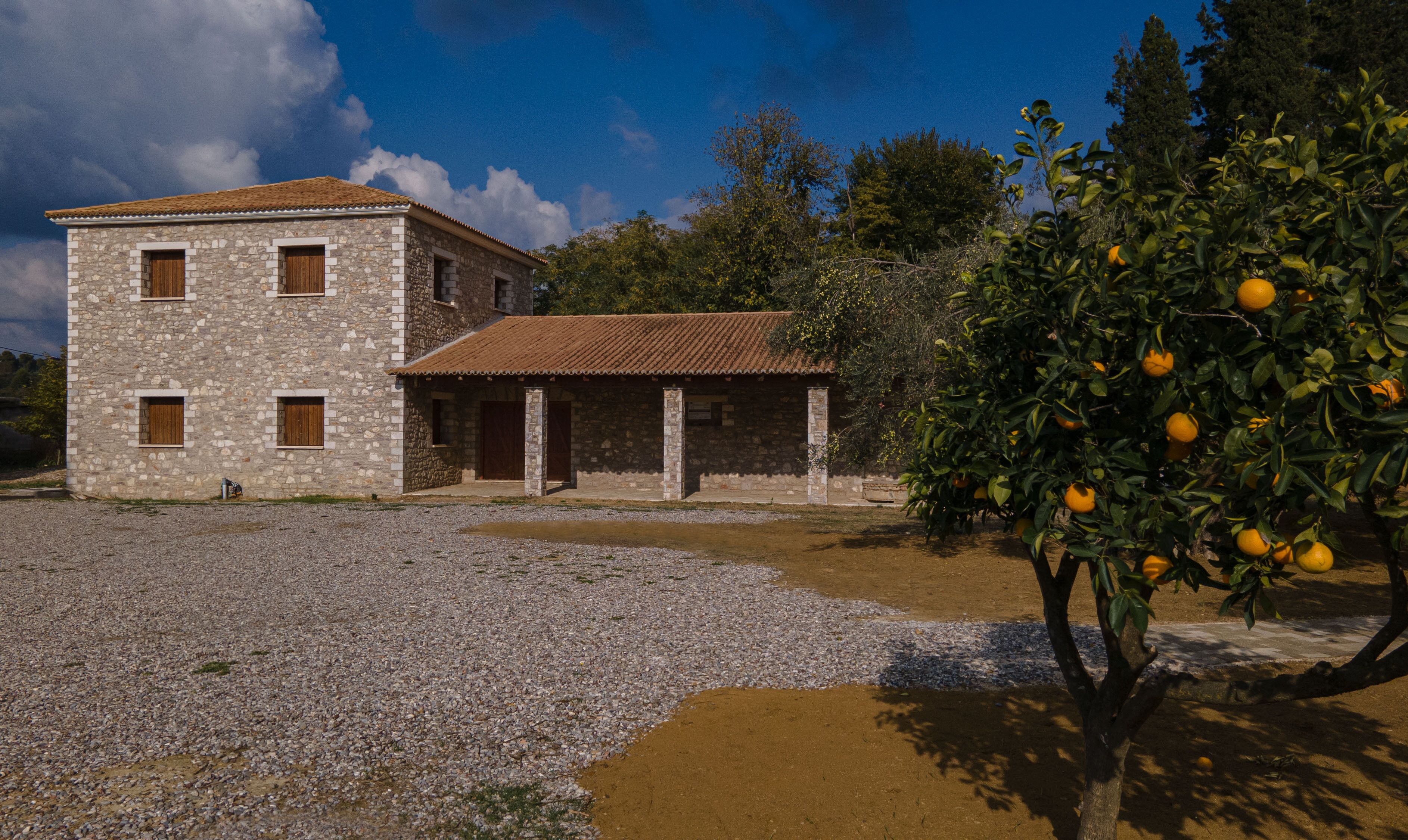 A stone building with a tiled roof, a gravel driveway, and an orange tree with fruit.
