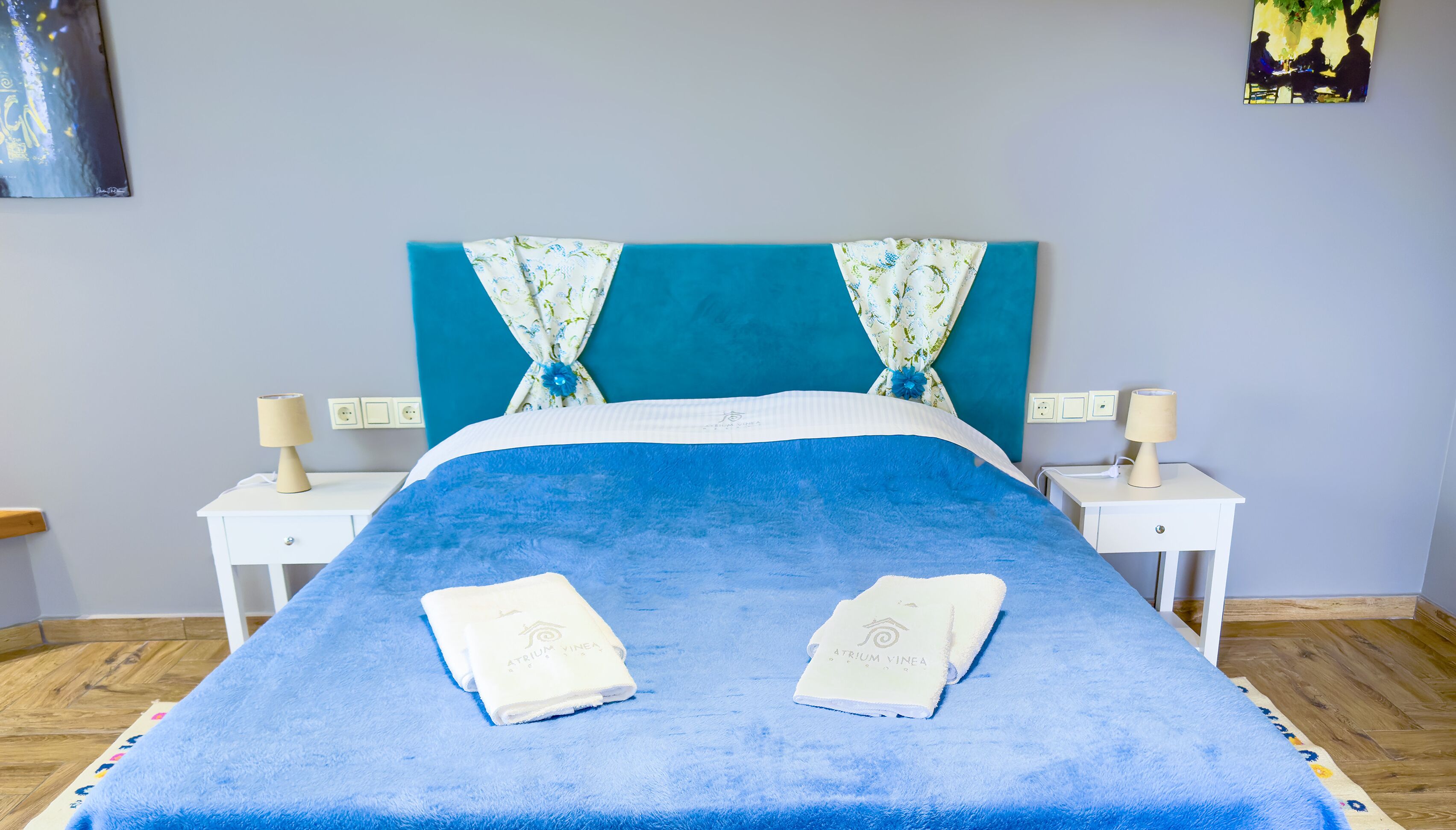 A neatly made bed with a blue blanket and two towels, white nightstands with lamps, and a painting on the wall.