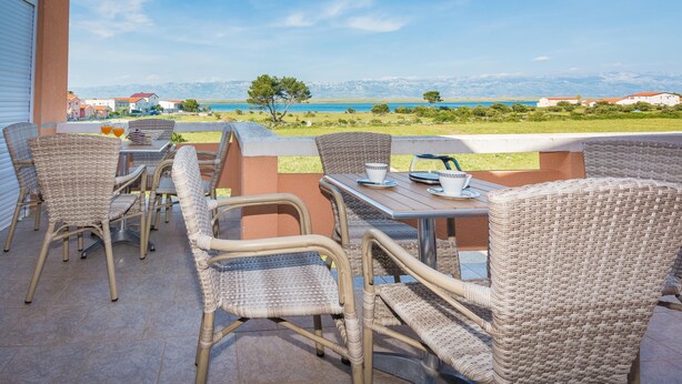Un patio al aire libre con mobiliario de mimbre, una mesa con tazas y una botella, y vista al mar y las montañas de fondo.