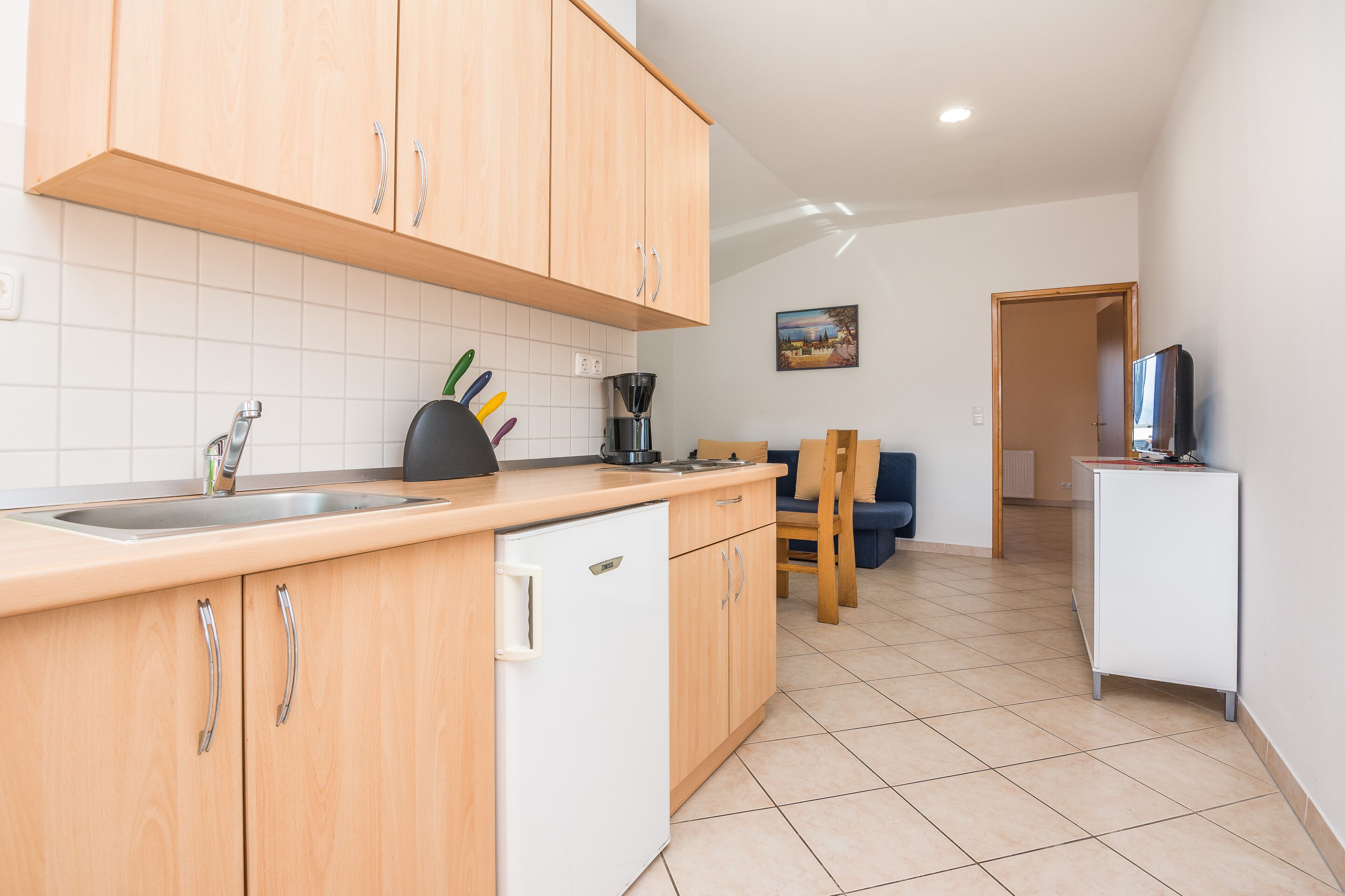 A kitchen with wooden cabinets, a sink, and a coffee maker. There is a chair and a television in the background.