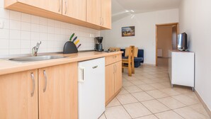 Una cocina con armarios de madera, un lavamanos y una cafetera. Hay una silla y un televisor al fondo.