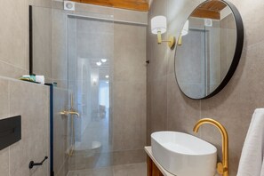 A modern bathroom with a glass shower enclosure, a white sink with a gold faucet, and a round mirror on the wall. - Casa Del Balsamo (Rethymno)