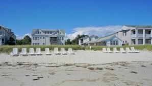 Ein Strandgrundstück mit mehreren Gebäuden, darunter ein Haus im Landhausstil und ein größeres Gebäude mit Balkon. Mehrere weiße Liegestühle stehen auf dem sandigen Strand vor den Gebäuden.