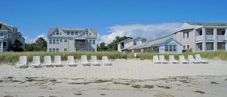 En fastighet vid stranden med flera byggnader, inklusive ett hus i stugstil och en större byggnad med en balkong. Framför byggnaderna, på den sandiga stranden, står flera vita solstolar uppställda.