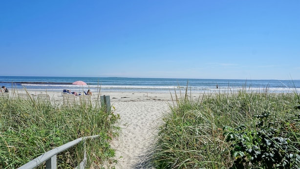 Ein Strandweg führt zu einem Sandstrand, wo einige Personen und ein Strandsonnenschirm zu sehen sind.