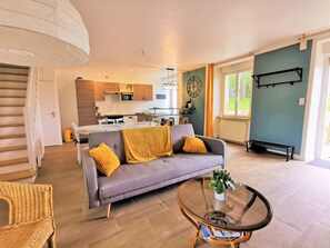 Ein modernes Wohnzimmer mit einem grauen Sofa, gelben Kissen, einem Glastisch, Korbstühlen und einer Küche im Hintergrund.