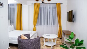 A hotel room with a bed, a small round table, a chair, a plant, and a TV mounted on the wall. - Santorina Home Suites (Kisii)