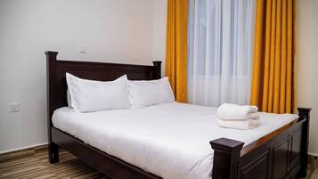Una cama bien hecha con sábanas y almohadas blancas, un cabecero de madera y una cortina amarilla ligeramente separada.