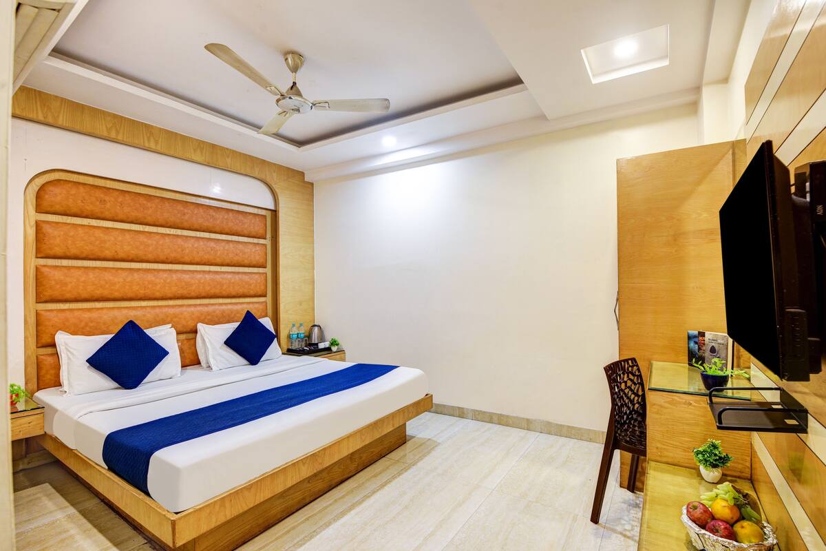 A modern hotel room with a wooden headboard, a bed with blue bedding, a ceiling fan, a small table with a plant, and a fruit basket.