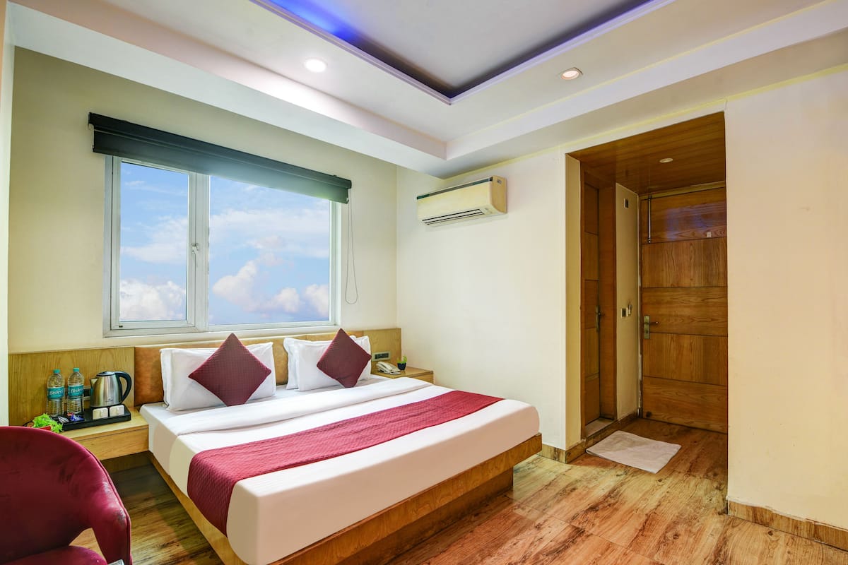 A hotel room with a large bed, a wooden floor, a window with a view, an air conditioning unit, and a door.