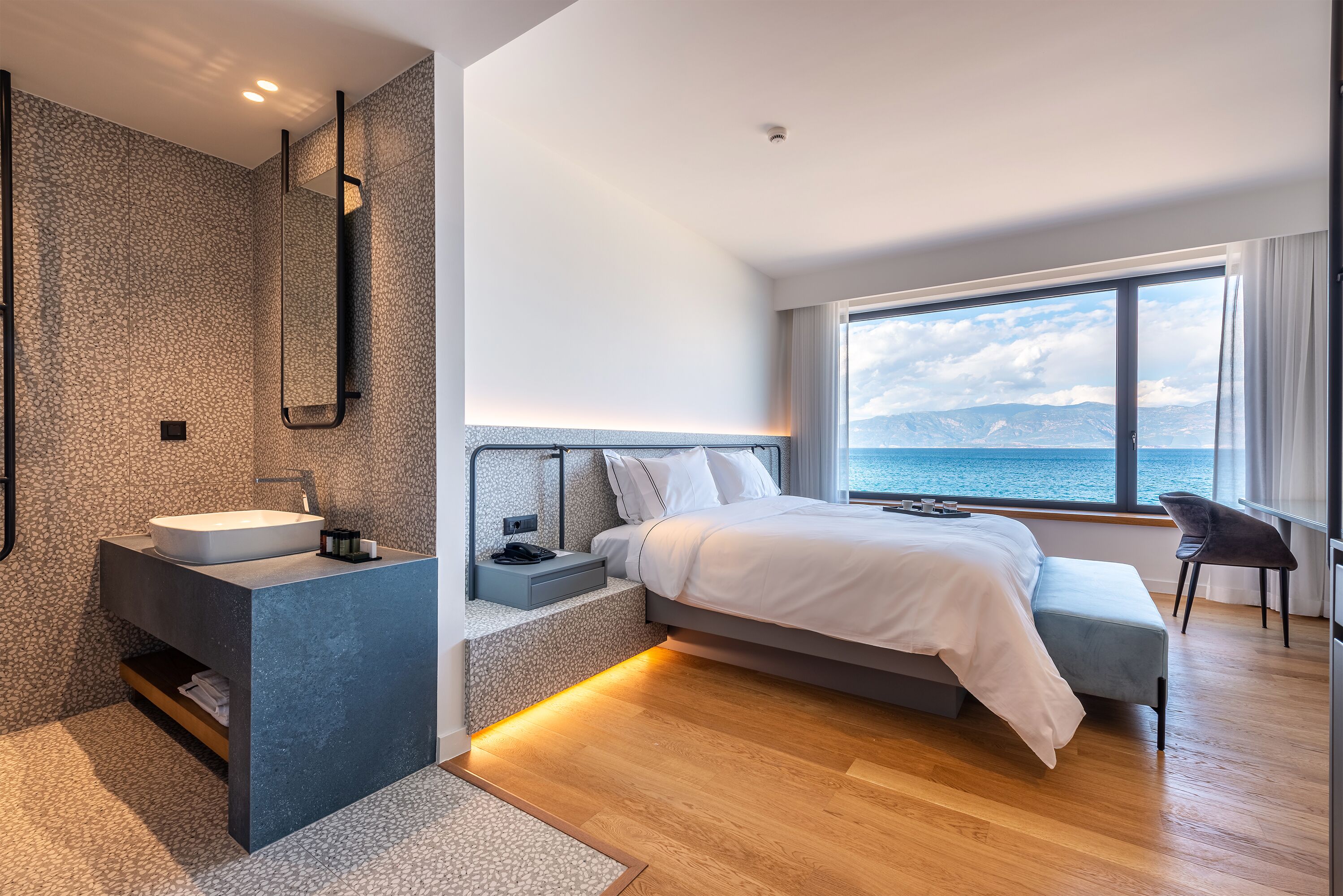 A modern bedroom with a large bed, a built-in vanity, and a view of the sea.
