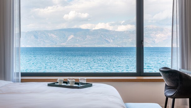 A hotel room with a large window offering a view of the sea and mountains. There is a bed with white linens, a tray with a cup and glasses, and a chair. - Amario Suites Hotel (Aigialeia)