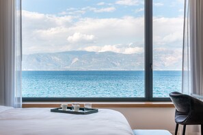 A hotel room with a large window offering a view of the sea and mountains. There is a bed with white linens, a tray with a cup and glasses, and a chair. - Amario Suites Hotel (Aigialeia)