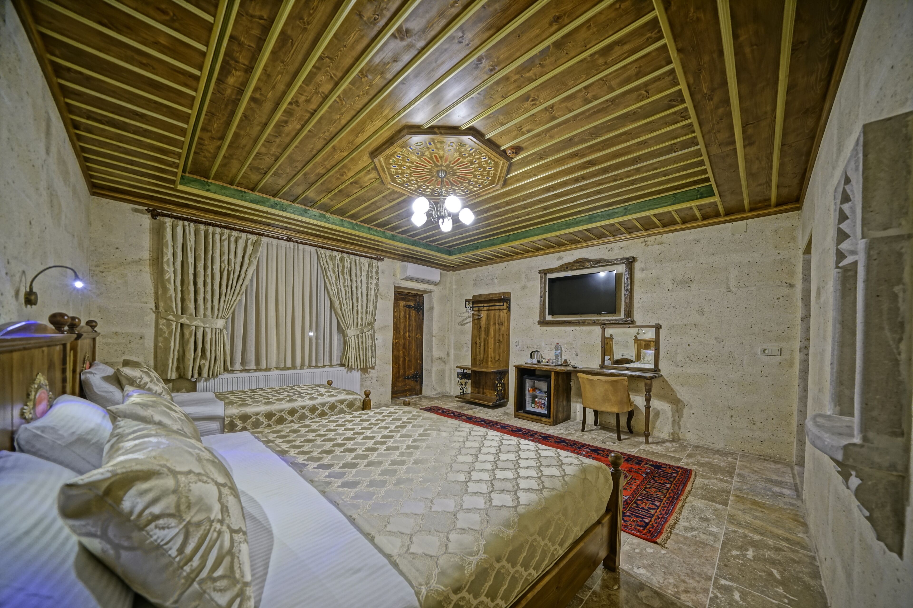 A room with wooden beams on the ceiling, a bed with a patterned bedspread, a small desk, and a television on the wall.