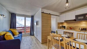 Een moderne woonkamer met een bank, een eettafel en stoelen, een keuken met houten kastjes, en uitzicht op een besneeuwde berg.
