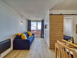A modern living room with a blue sofa, wooden flooring, and a view of snow-capped mountains through a sliding glass door. - Altineige by Interhome (Val Thorens)