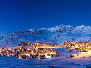 Une ville de montagne enneigée, au crépuscule, avec des bâtiments illuminés et une piste de ski.