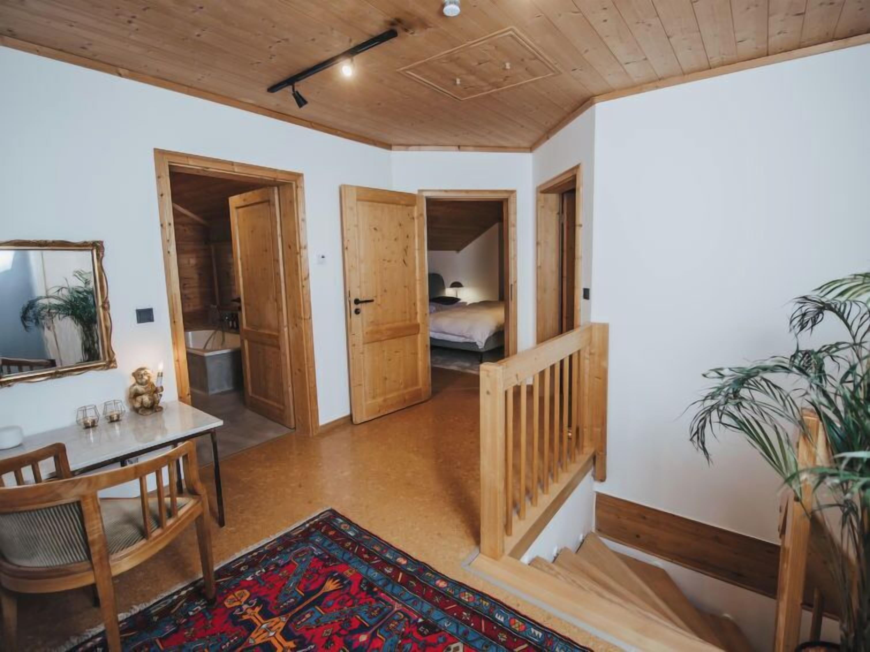 Ein gemütliches Zimmer mit Holzbalkendecke, einem Esstisch mit Stühlen, einem farbenfrohen Teppich und einer Treppe, die in ein weiteres Zimmer führt.