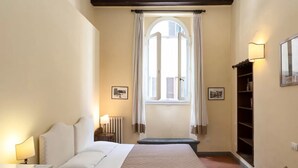 Une chambre à coucher comprenant un lit, une table de chevet, un radiateur, une fenêtre avec des rideaux, un banc et une bibliothèque.