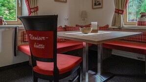 Zona pranzo con tavolo, sedie e una panca. Due quadri appesi al muro, una finestra con tende e un termosifone.