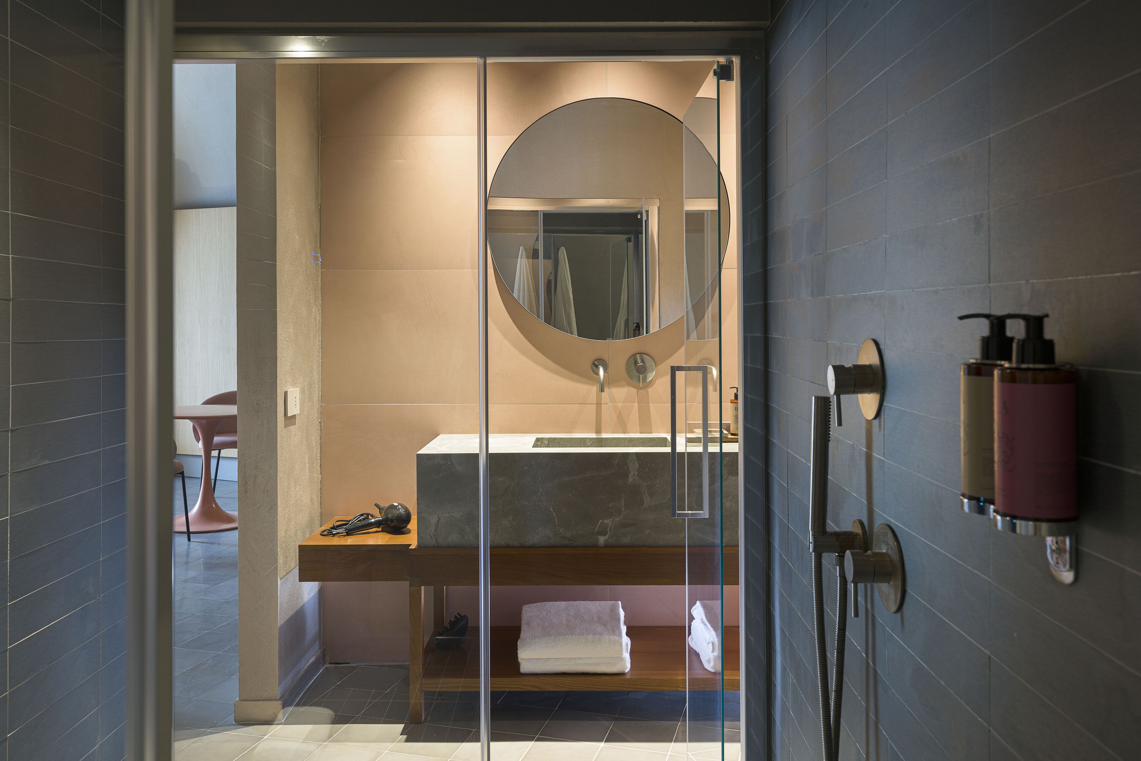 A modern bathroom with a glass-enclosed shower, a round mirror, a marble sink, and a towel rack.