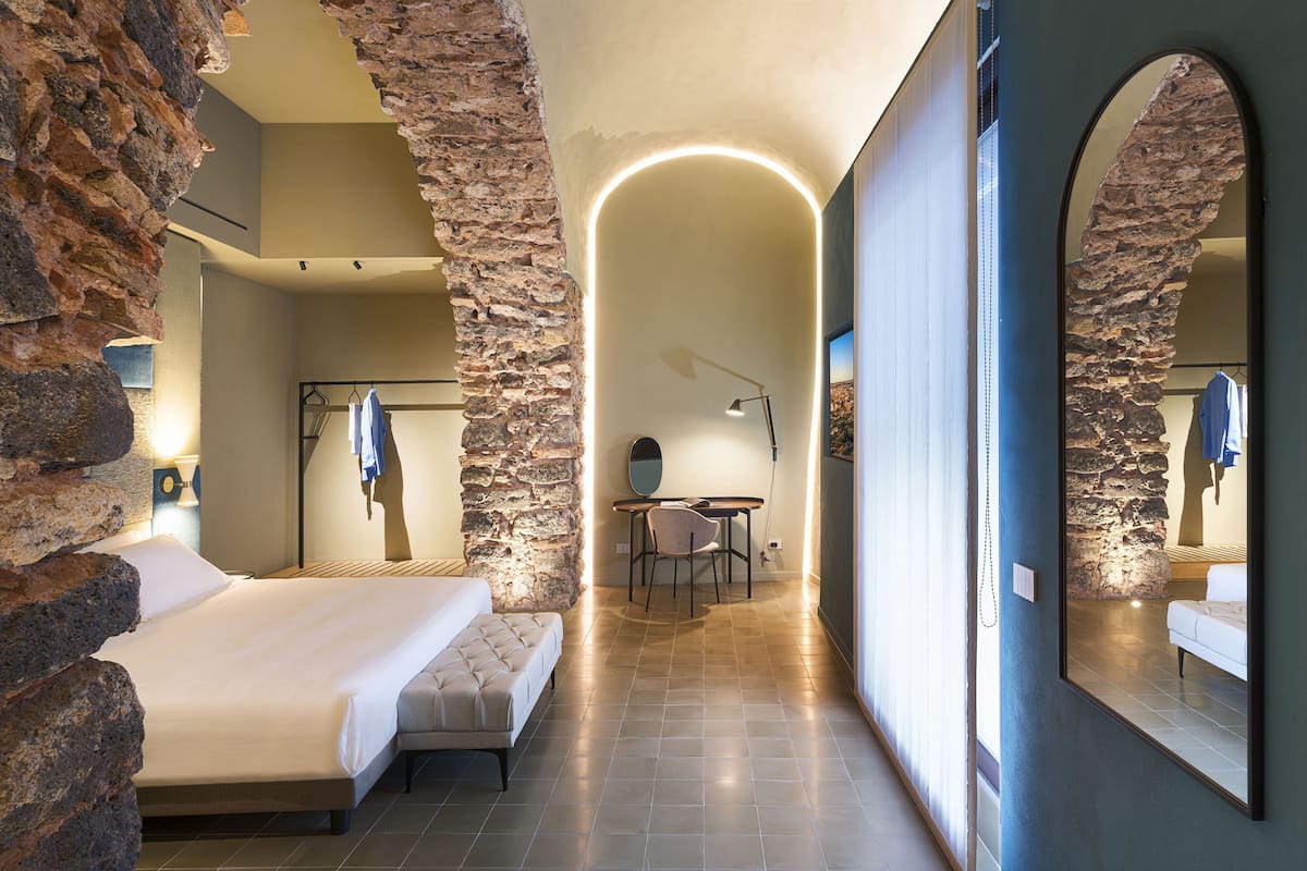 A hotel room with a bed, a desk, a chair, a mirror, and stone walls.