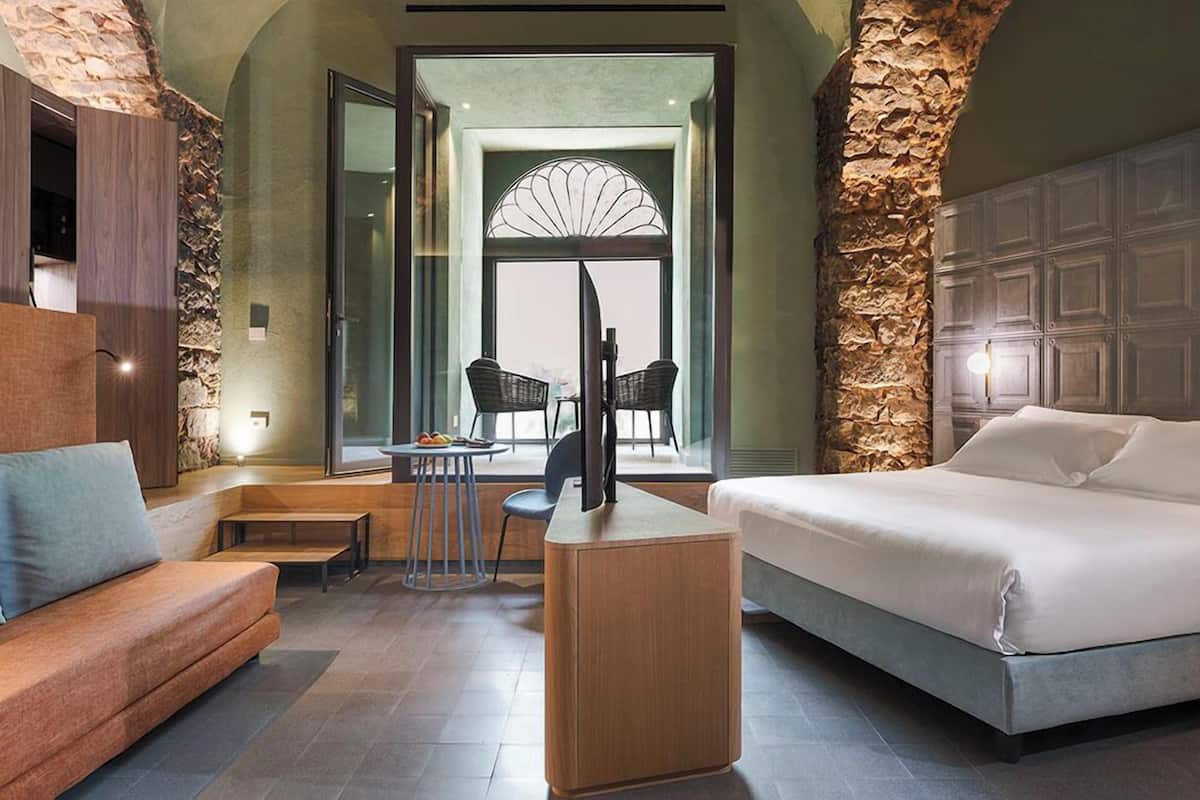 A modern hotel room with a stone accent wall, a large bed, a sofa, a small table with chairs, and a TV.