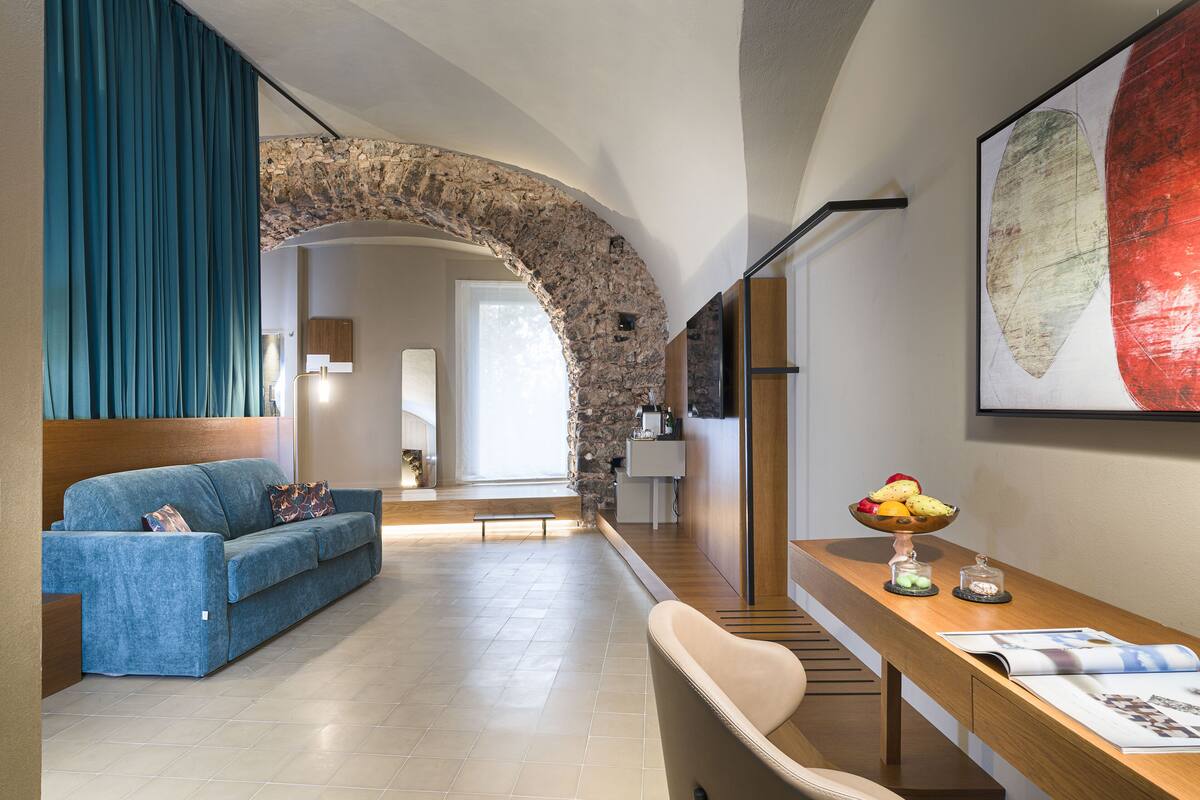 A modern living room with a stone archway, a blue sofa, a wooden dining table with a fruit bowl, and a large abstract painting.