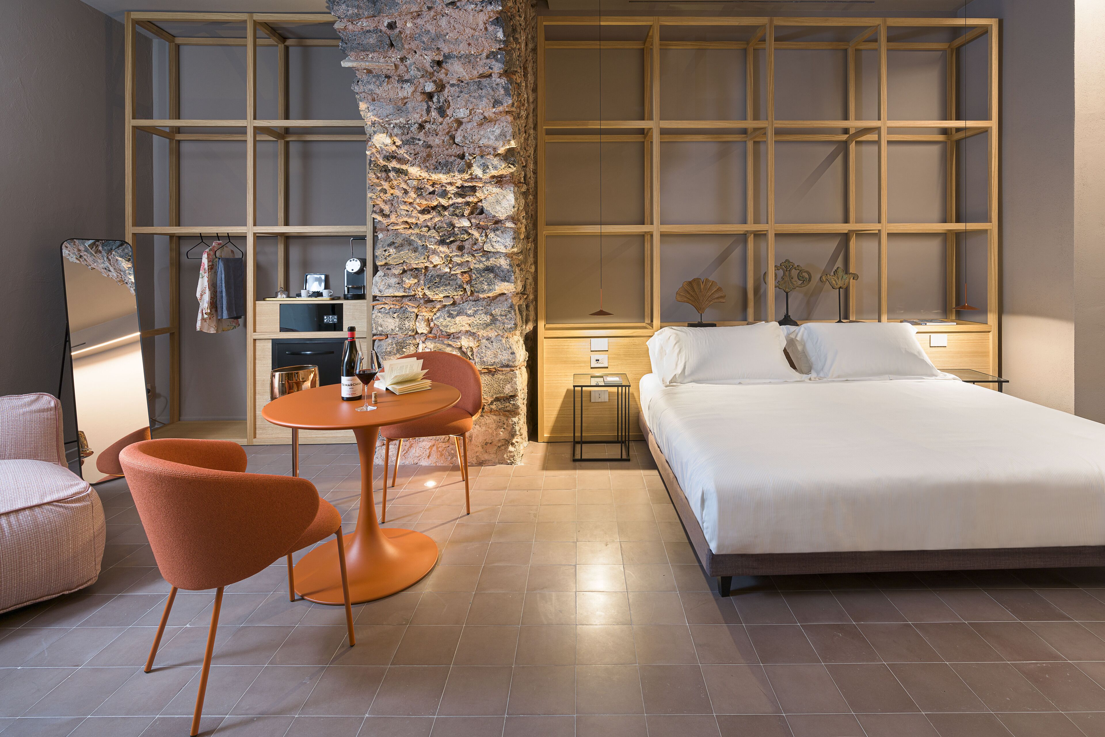A modern hotel room with a large bed, a small round table with two chairs, a stone accent wall, and a wooden shelving unit.