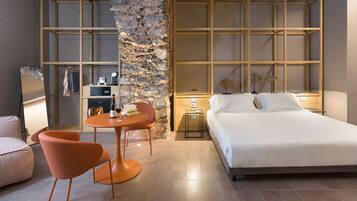 A modern hotel room with a large bed, a small round table with two chairs, a stone accent wall, and a wooden shelving unit.
