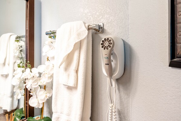 A bathroom with a towel rack holding white towels, a hairdryer mounted on the wall, and white orchids in a vase.