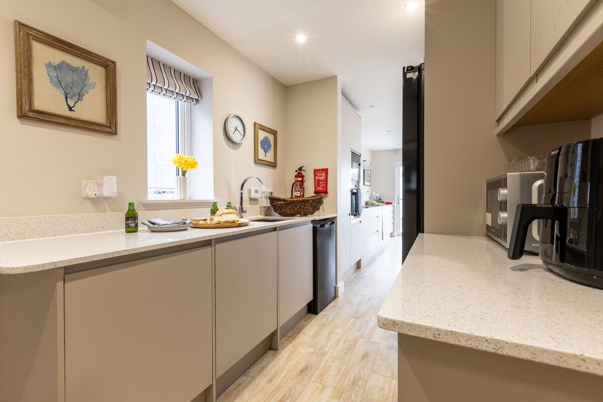 A modern kitchen with a countertop, a sink, a dishwasher, and a refrigerator. There is a window with a view of the outside, a clock on the wall, and a framed picture above the sink.
