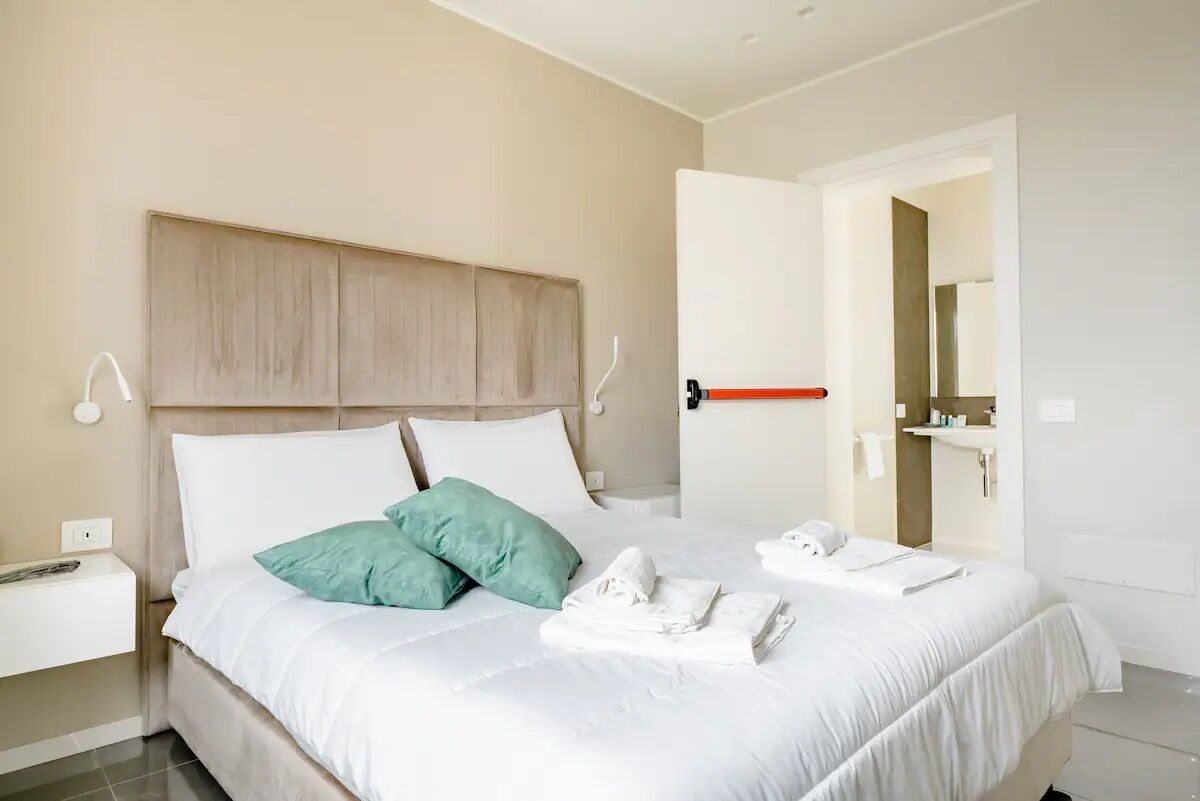 A neatly made bed with white bedding and two green pillows, a bedside table with a lamp, and a bathroom visible through an open door.
