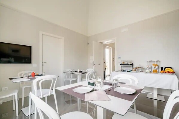 A modern dining area with white chairs and tables, a flat-screen TV mounted on the wall, and a breakfast setup with fruits and pastries.